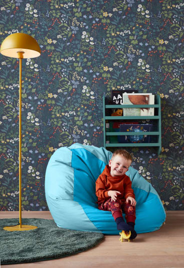 Ein kleines Kind sitzt auf einem blauen Sitzsack vor einer blumengeschmückten Wand. Links steht eine gelbe Stehlampe, und hinter dem Kind ist ein grünes Wandregal mit Büchern zu sehen. Ein grüner Teppich und ein Holzboden sind ebenfalls zu sehen.