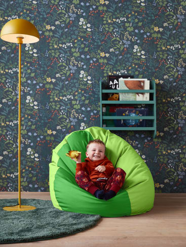 Ein kleines Kind sitzt lächelnd auf einem hellgrünen Sitzsack und hält ein gelbes Spielzeug. Im Hintergrund befinden sich eine Blumentapete, ein grüner Teppich, eine gelbe Stehlampe und ein kleines Bücherregal mit Büchern an der Wand.
