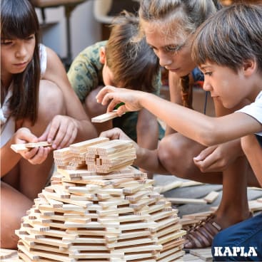 Vier Kinder bauen eine große, kuppelförmige Struktur aus rechteckigen Kapla-Holzbrettern. Sie sind konzentriert, arbeiten zusammen und fügen die Teile sorgfältig zusammen. Das Kapla-Logo ist in der unteren rechten Ecke zu sehen.