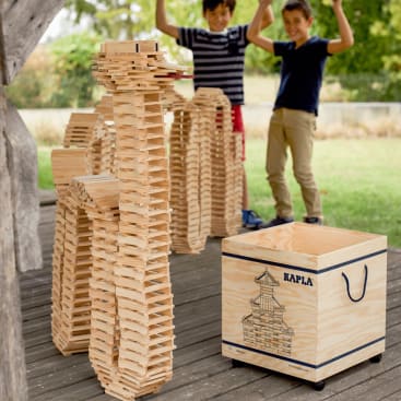 Zwei Kinder stehen hinter großen Tierskulpturen, die aus Kapla-Holzbrettern gebaut sind. Davor steht auf dem Holzboden eine Kapla-Aufbewahrungsbox mit einer Burgdarstellung. Der Schauplatz scheint ein überdachter Außenbereich zu sein.