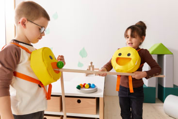 Zwei Kinder in gelben Emoji-Kostümen spielen ein Spiel. Der Junge versucht, einen grünen Ball mit dem Mund seines Kostüms aufzuheben, während das Mädchen einen Stock hält. Ein Teller mit bunten Bällen steht auf dem Tisch hinter ihnen.