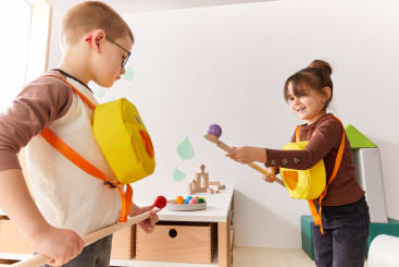 Zwei Kinder spielen zusammen in einem Haus. Beide tragen einen gelben Rucksack und halten einen Holzstab mit einem Ball in der Hand. Sie stehen sich gegenüber, und im Hintergrund steht ein Tisch mit Spielzeug.