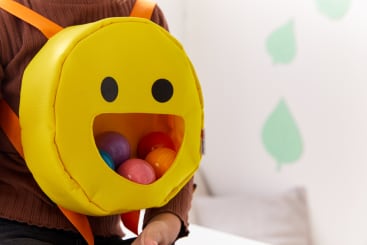Eine Person hält einen gelben, runden Rucksack in Form eines lächelnden Gesichts mit einem großen offenen Mund, der mit bunten Plastikbällen gefüllt ist. Die Person trägt ein braunes Oberteil. An der Wand im Hintergrund befinden sich grüne Blätter.