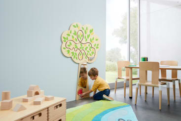 Ein kleines Kind kniet auf einem Teppich in einem modernen Klassenzimmer und spielt mit einem roten Spielzeug auf einem niedrigen, an der Wand befestigten Spiegel, der auch eine hölzerne Dekoration in Form eines Baumes mit aufgemalten grünen Blättern aufweist.