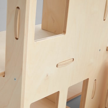Nahaufnahme von hellen Holzplatten mit ausgeschnittenen Griffen und rechteckigen Öffnungen, die durch sichtbare Schrauben verbunden sind. Die Oberfläche weist eine glatte, natürliche Holzmaserung auf.
