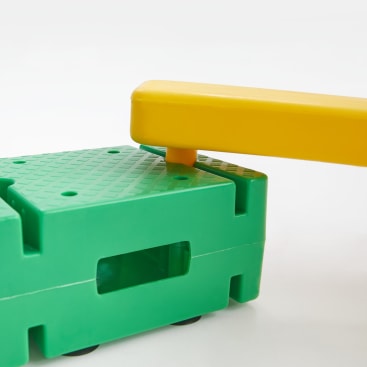Nahaufnahme eines gelben Kunststoffbalkens, der mit einem grünen Kunststoffsockel mit strukturierter Oberfläche und rechteckigem Loch verbunden ist; beide Teile sind wahrscheinlich Teil eines modularen Spielzeugs oder Bausatzes.