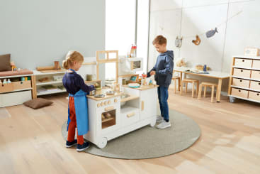 Zwei Kinder spielen mit einer Holzspielzeugküche in einem hellen Klassenzimmer mit Holzmöbeln, Regalen und Spielzeug. Ein Kind trägt eine Schürze, während beide so tun, als würden sie kochen. Der Raum hat helle Holzböden und neutrale Wände.