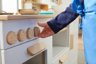 Eine Person in einer blauen Schürze dreht einen Knopf an einem weißen Spielzeugküchenherd mit Holzteilen. Auf der Arbeitsplatte im Hintergrund sind weitere Küchenutensilien, wie z. B. eine Pfanne, zu sehen.