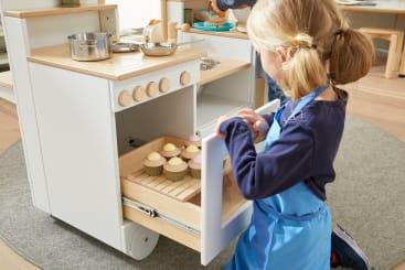 Ein junges Mädchen in einer blauen Schürze öffnet einen Spielzeugofen und legt ein Tablett mit Spielzeug-Cupcakes mit pastellfarbenem Zuckerguss in einer Spielküche frei. Töpfe und Pfannen sind auf der Herdplatte darüber zu sehen.