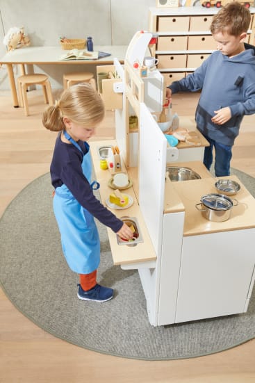 Zwei kleine Kinder spielen mit einem Holzspielzeug-Küchenset. Ein Kind trägt eine blaue Schürze und legt Spielzeugessen in einen Ofen, während das andere in der Nähe steht und Gegenstände auf einer Arbeitsplatte anordnet. Im Hintergrund ist ein Klassenzimmer zu sehen.
