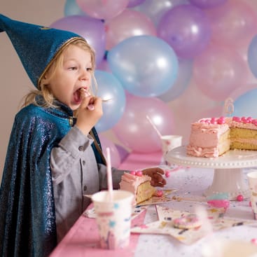 Ein Kind mit blauem Zaubererhut und Umhang isst ein Stück rosa Gugelhupf an einem dekorierten Tisch mit Luftballons und Partyzubehör. Der Kuchen steht auf einem weißen Ständer mit einer Kerze mit der Nummer sechs darauf.