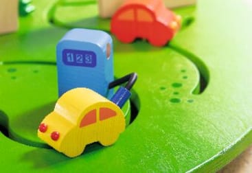 Ein gelbes Spielzeugauto aus Holz ist durch einen Schlauch mit einer blauen Benzinpumpe auf einer grünen Spielmatte verbunden. Im Hintergrund ist ein rotes Spielzeugauto zu sehen.