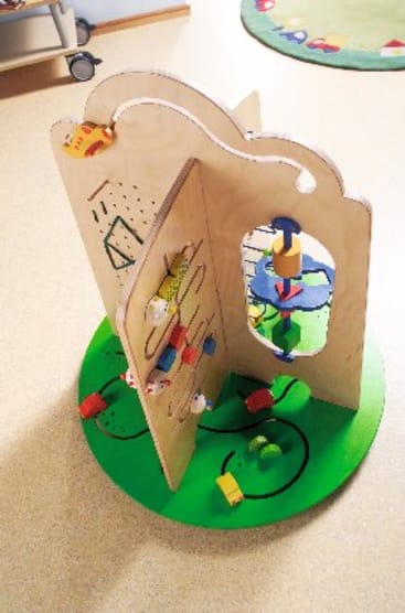 Aktivitätszentrum aus Holz für Kinder mit interaktiven Elementen wie Perlen, Zahnrädern und einem Spiegel auf einem grünen runden Sockel auf einem beigen Boden. Im Hintergrund sind ein Teil eines Teppichs und Möbel zu sehen.