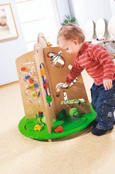 Ein kleines Kind in einem rot gestreiften Hemd und einer blauen Jeans spielt in einem hellen, mit Teppich ausgelegten Raum mit einem großen hölzernen Spielbrett mit verschiedenen Perlenlabyrinthen und bunten beweglichen Teilen.