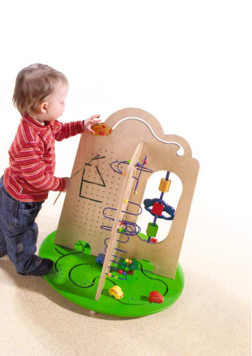 Ein kleines Kind steht und spielt mit einem großen hölzernen Spielbrett mit Perlenlabyrinthen, sich drehenden Zahnrädern und verschiedenen bunten interaktiven Elementen auf einem grünen Sockel.