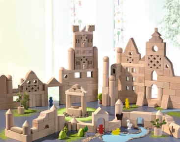 Mehrere Holzbausteine werden auf einer Spielfläche zu schlossähnlichen Gebilden zusammengesetzt, wobei kleine bunte Figuren, Scheinpflanzen und ein blauer Teich Teil der Szene sind.