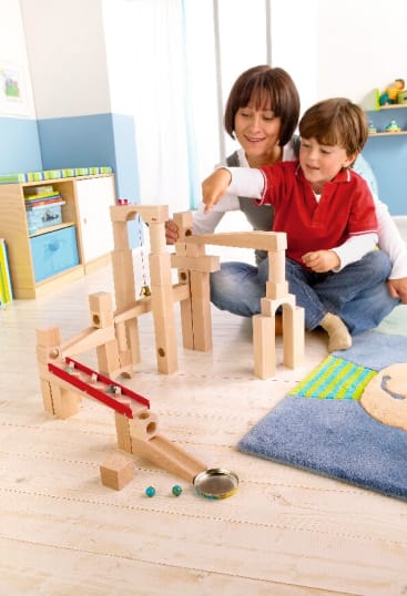 Eine Frau und ein Junge sitzen in einem hell erleuchteten Raum auf dem Boden und bauen mit Holzklötzen und Rampen eine Murmelbahn. Einige Murmeln liegen auf dem Boden neben dem Bauwerk.